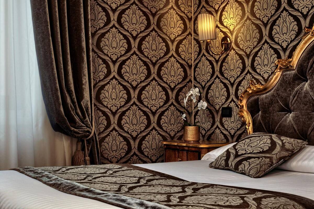 Une chambre à coucher luxueuse avec un papier peint à motifs, une tête de lit capitonnée, une table de chevet avec une lampe et un vase décoratif contenant une orchidée.
