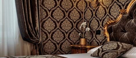 A luxurious bedroom with a patterned wallpaper, a tufted headboard, a bedside table with a lamp, and a decorative vase with an orchid.