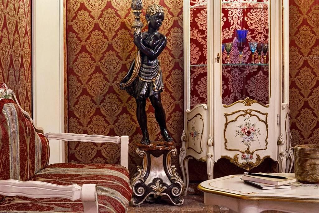 Une pièce avec un papier peint à motifs, une statue décorative, un meuble blanc aux portes vitrées et un canapé à rayures.