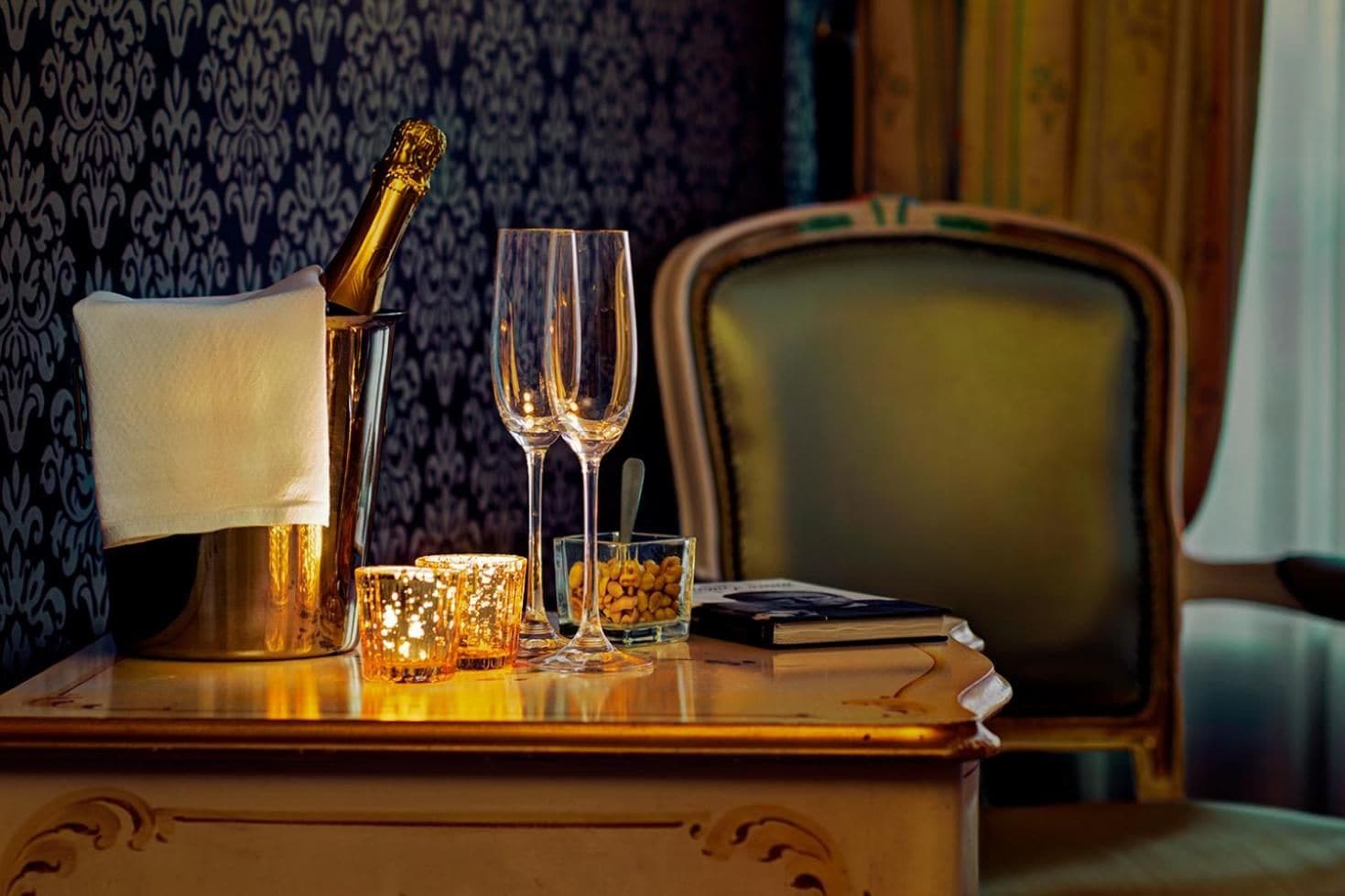 A wooden table with a champagne bottle, two glasses, a candle, and a bowl of nuts.