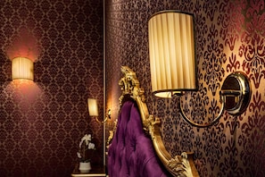 Una habitación con papel tapiz ornamentado, un cabecero tapizado en morado y lámparas fijadas a la pared.