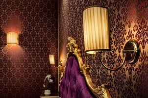 A room with ornate wallpaper, a purple upholstered headboard, and wall-mounted lamps.