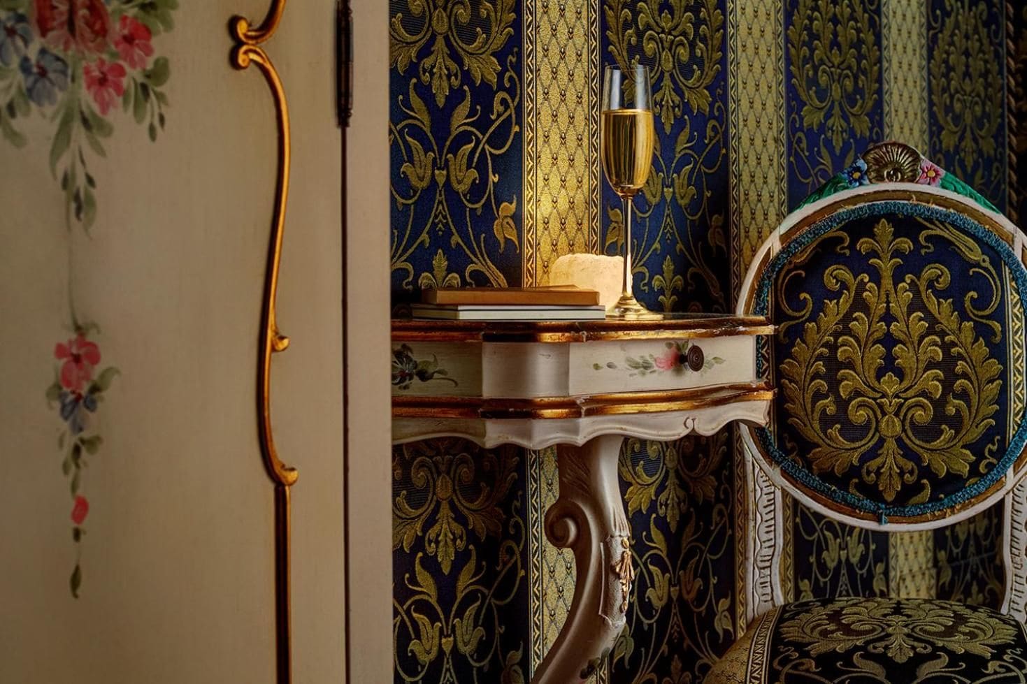 Quarto com uma cadeira decorativa, uma mesinha com um livro e um copo de vinho, e uma parede com papel de parede floral.