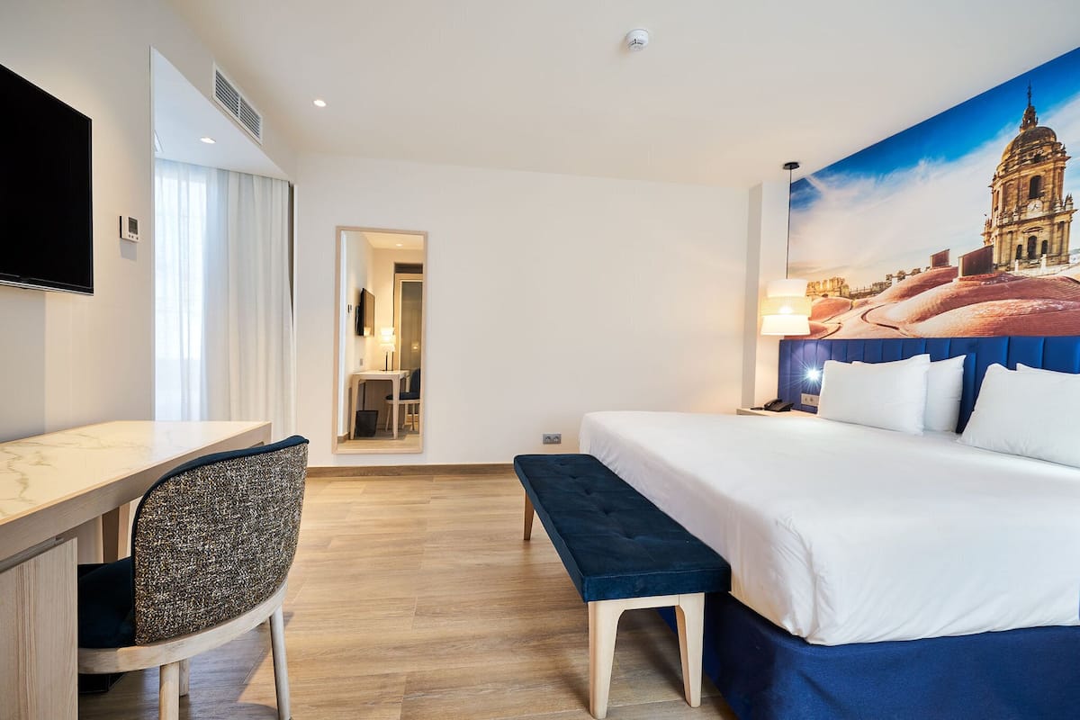 Ein Hotelzimmer mit Bett, Schreibtisch, Stuhl, einer Bank, einem Fernseher und einem großen Fenster.