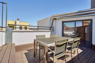Una terrazza sul tetto con pavimento in legno, un tavolo con sedie e una porta vetrata che dà su un'area interna.