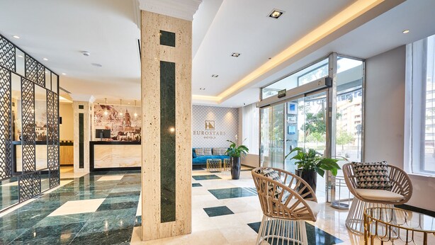 Un hall de hotel con suelo a cuadros, sillas de mimbre, un mostrador de recepción y ventanas amplias.