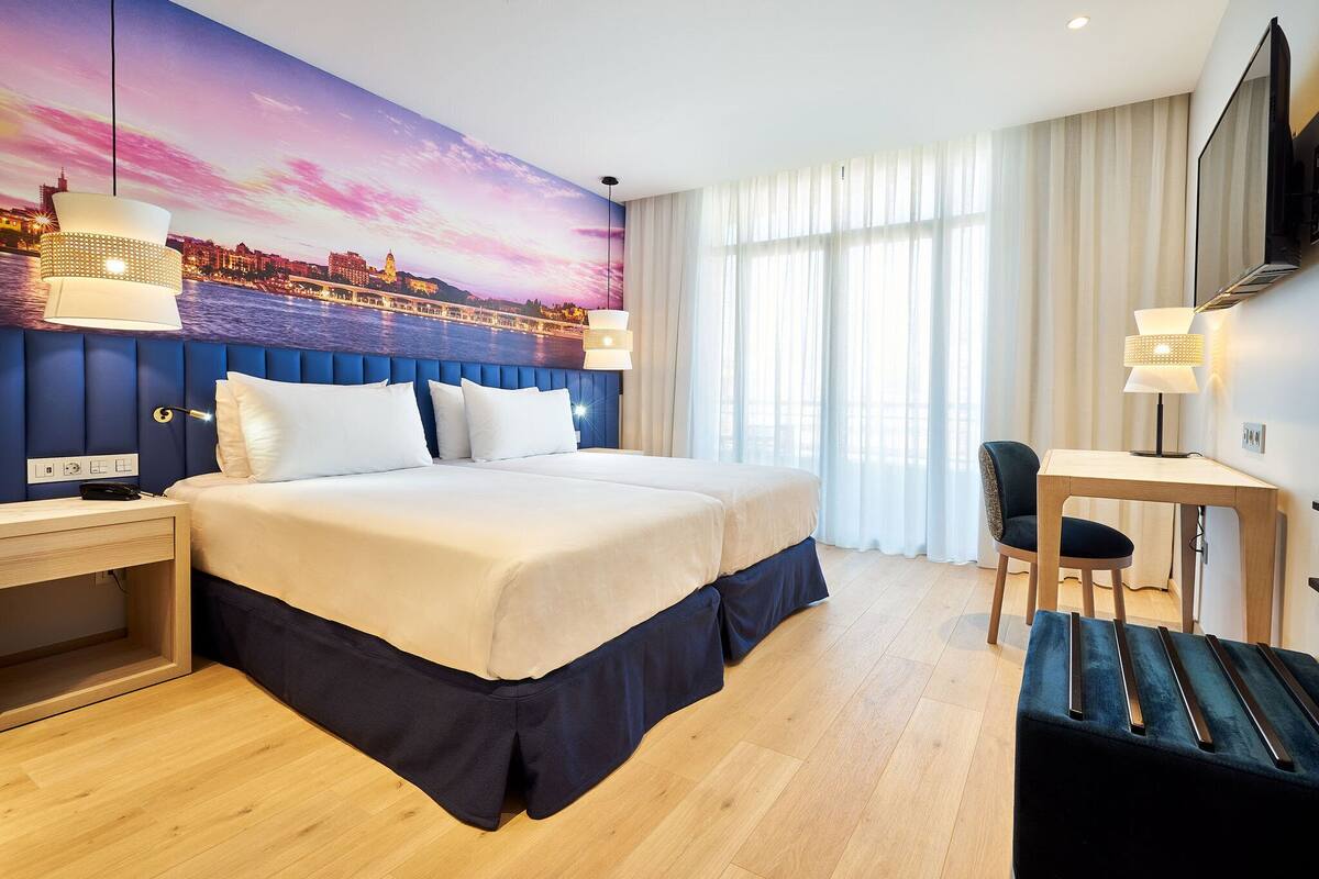 Ein Hotelzimmer mit einem großen Bett, einem Schreibtisch und einem Stuhl. An der Wand hängt ein Gemälde und es gibt ein Fenster mit Vorhängen.