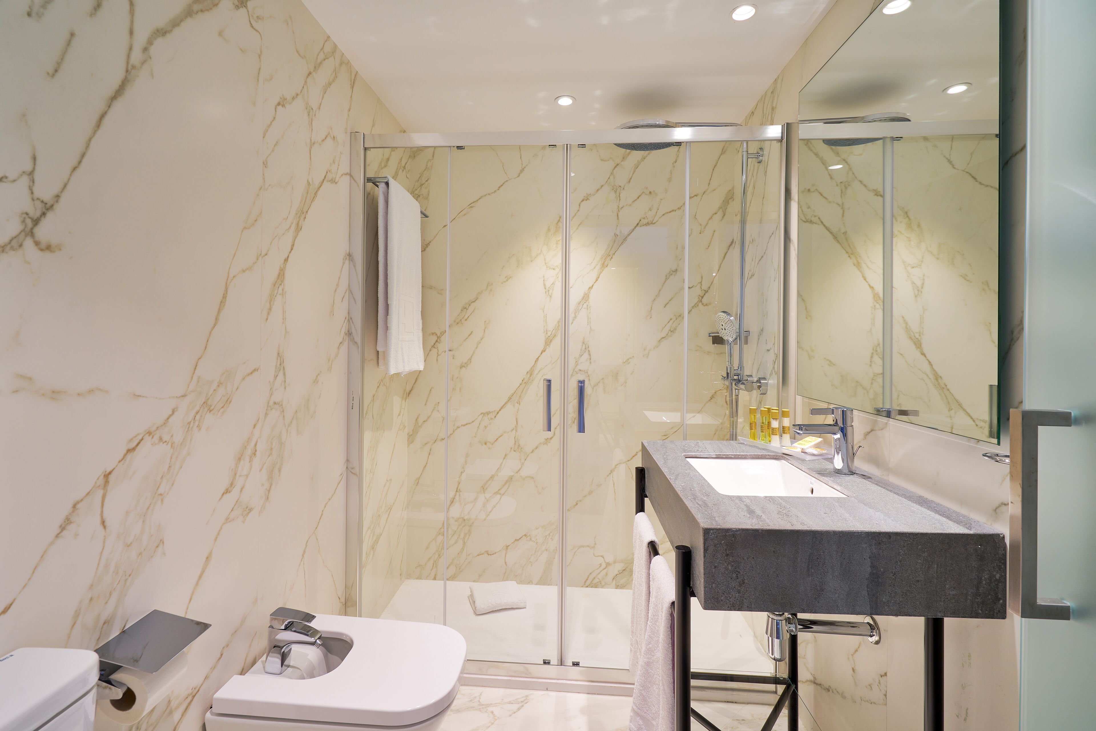 A modern bathroom with a marble wall, a glass-enclosed shower, a white toilet, and a sink with a dark countertop.