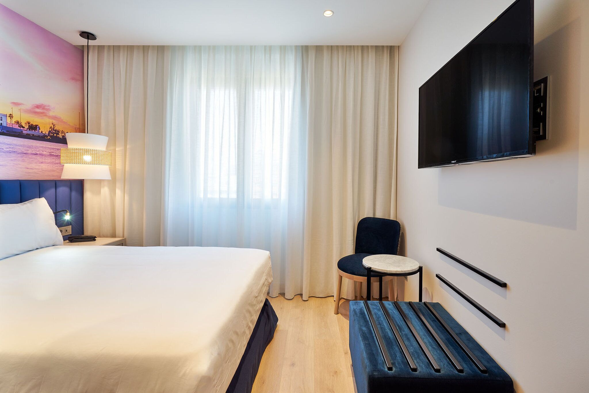 Habitación de hotel con cama, escritorio, silla, televisión y ventana con cortinas.