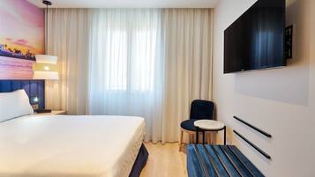 Habitación de hotel con cama, escritorio, silla, televisión y ventana con cortinas.