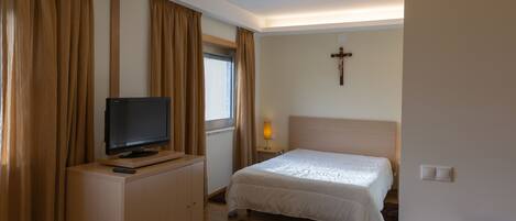 Un dormitorio con una cama, un televisor sobre un mueble, una cruz en la pared y una ventana con cortinas.