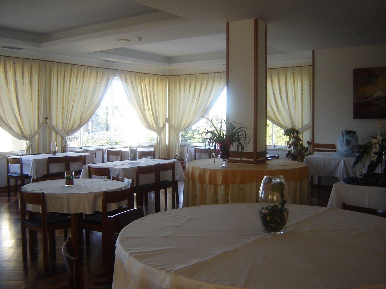 A restaurant with round tables and chairs, large windows with curtains, and a painting on the wall.