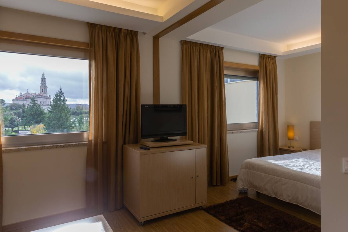 Ein Hotelzimmer mit Bett, Fernseher auf einer Kommode, einem Fenster mit Vorhängen und Blick auf ein Gebäude.