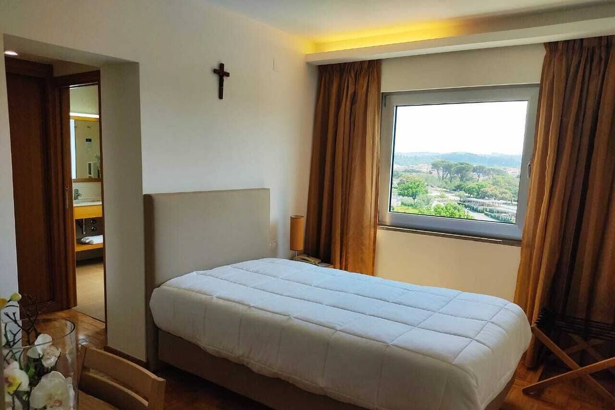 Ein Schlafzimmer mit einem Bett, einem Kreuz an der Wand, einem Fenster mit Vorhängen und Blick ins Grüne.