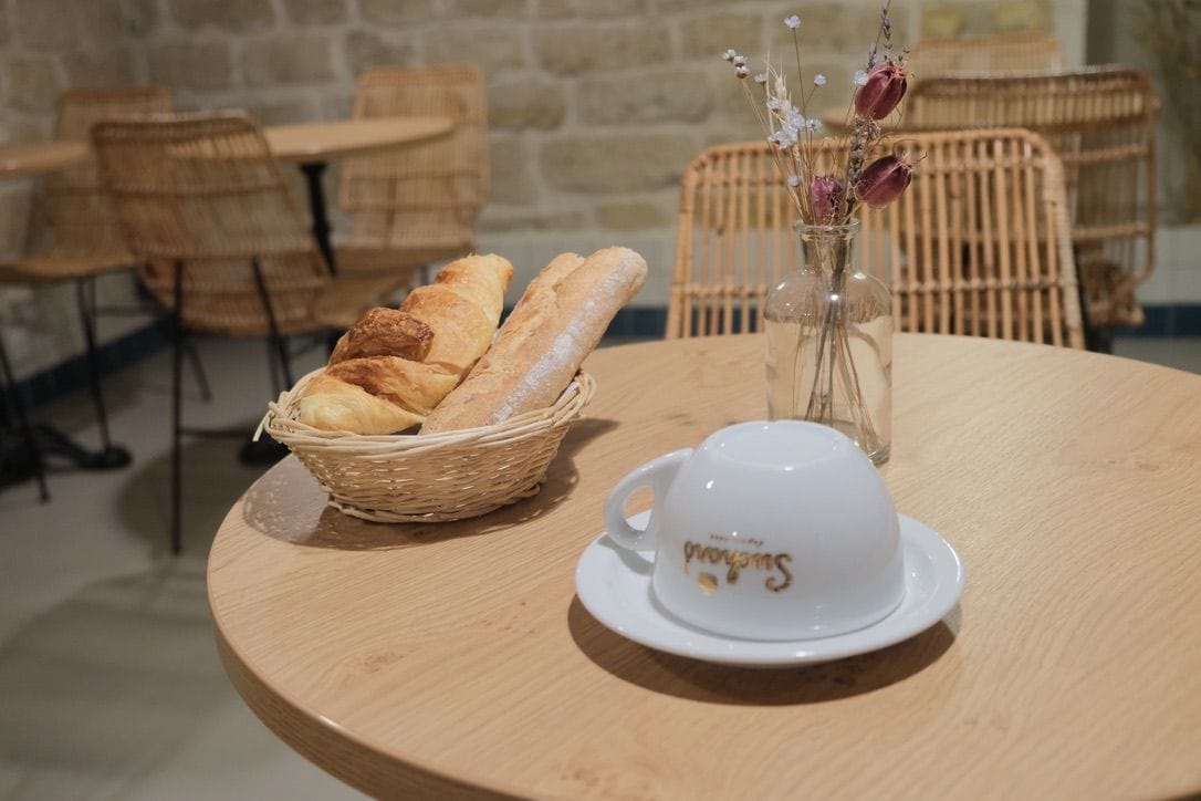 Una mesa redonda de madera con una cesta de pan, una taza blanca y un platillo, y un florero con flores secas.