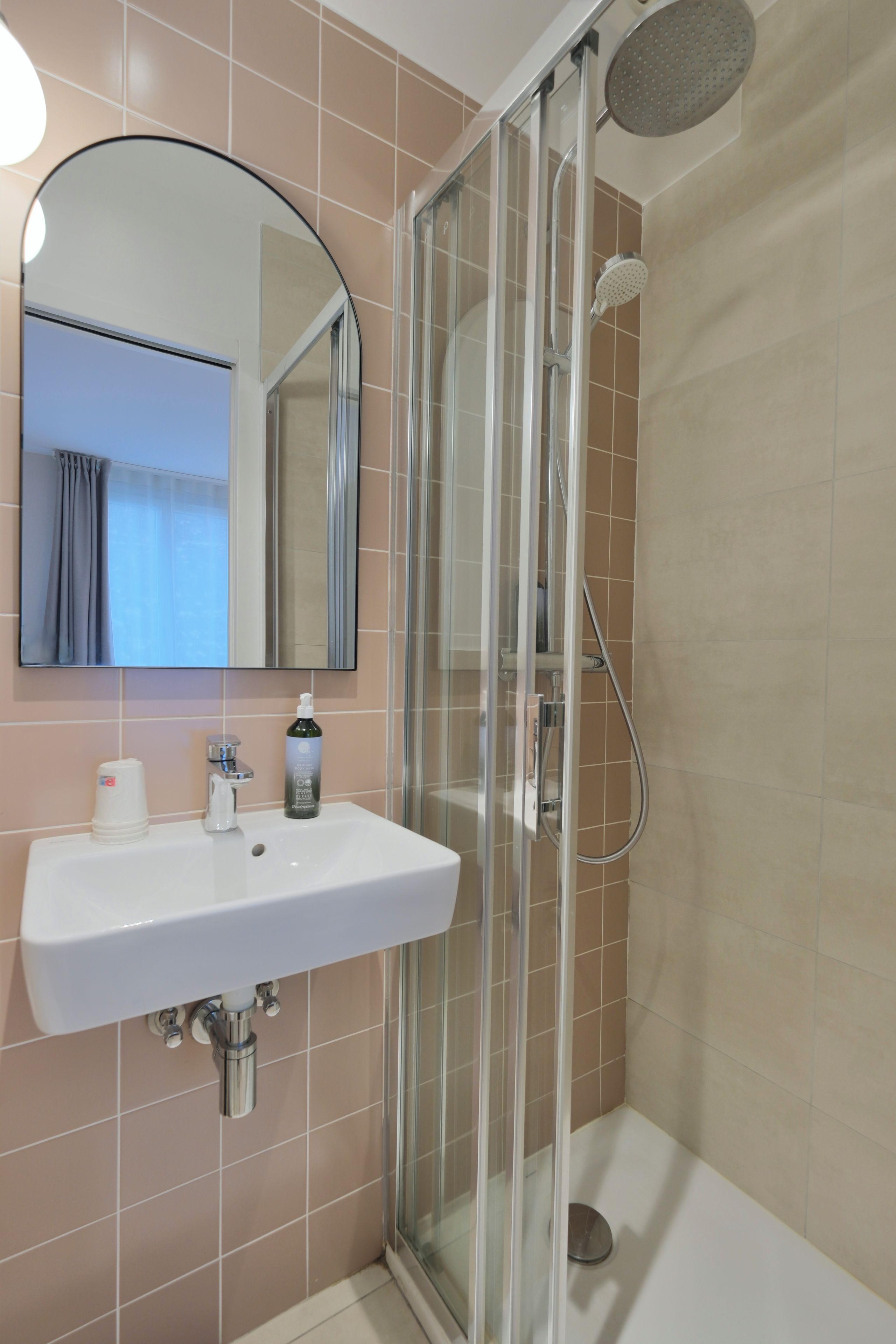 A bathroom with a glass-enclosed shower, a sink with a mirror above it, and a bottle of shampoo on the sink.