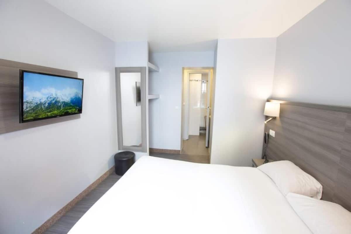 Ein Hotelzimmer mit Bett, einem an der Wand befestigten Fernseher, einem Nachttisch und einer Tür zu einem Badezimmer.