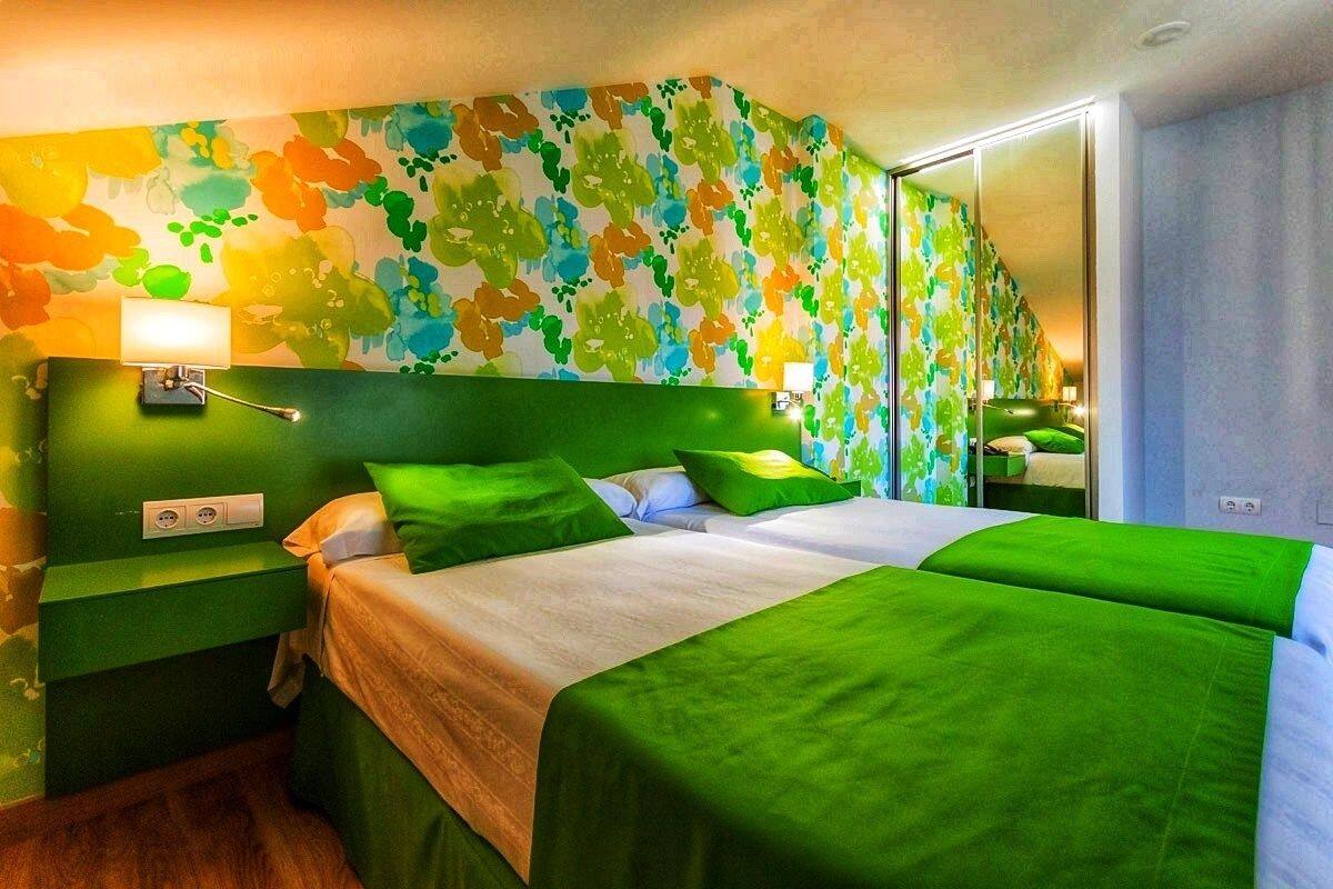 A hotel room with a green color scheme, a large bed, a nightstand, a mirror, and a wall with a floral pattern.