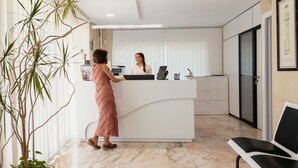 A reception desk with a woman standing behind it, a waiting area with chairs, and a large plant to the left. - Apartamentos Vértice Bib Rambla (Seville)