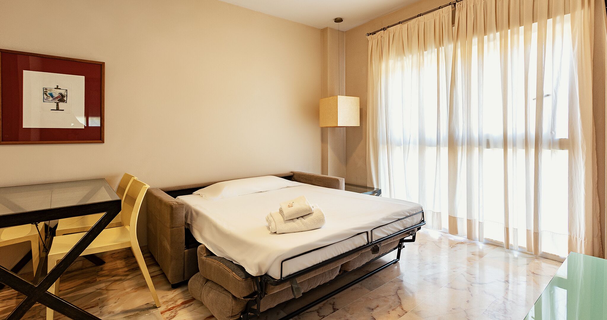 A hotel room with a bed, a desk, a chair, a lamp, and a window with curtains.
