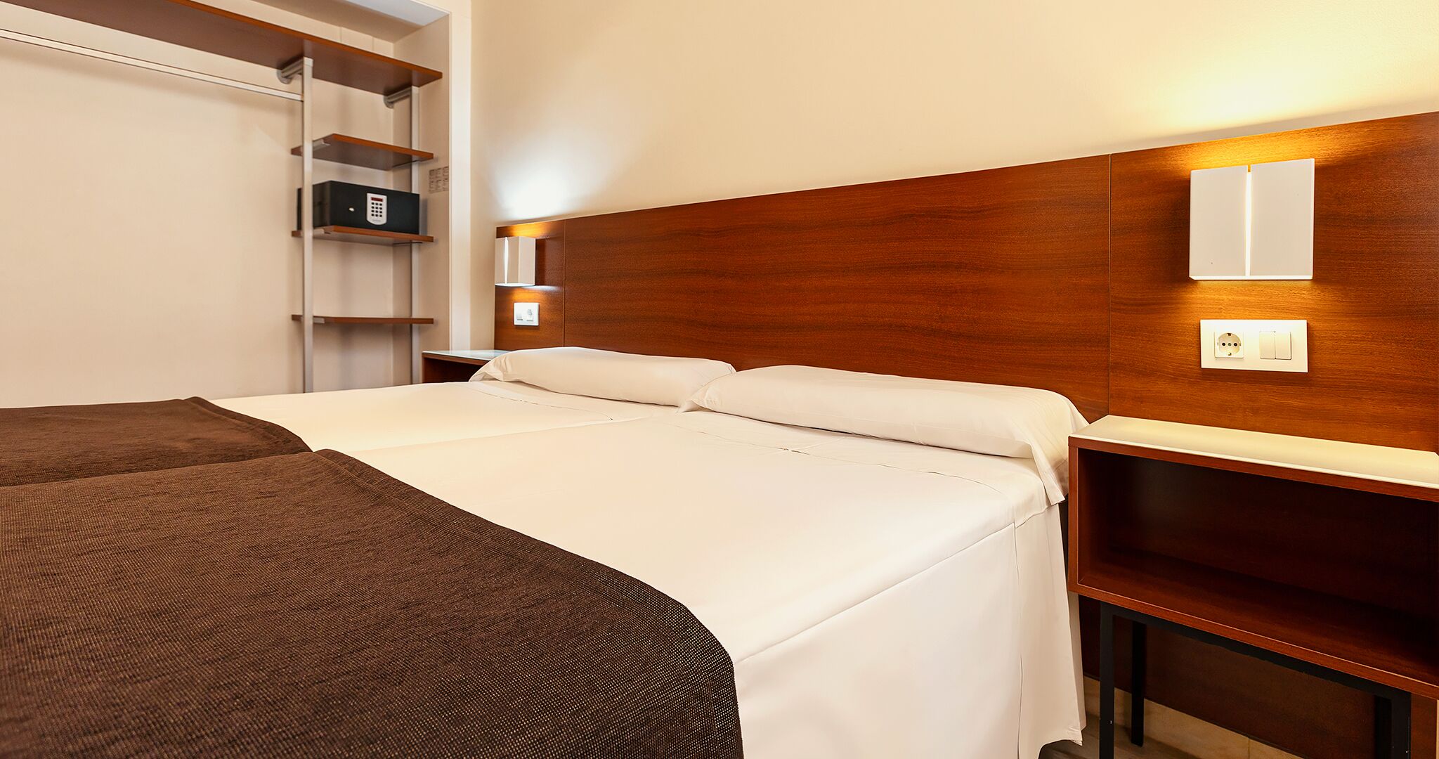 Een hotelkamer met een groot bed, een nachtkastje, een houten hoofdbord, een plank met een kluisje en een wandlamp.