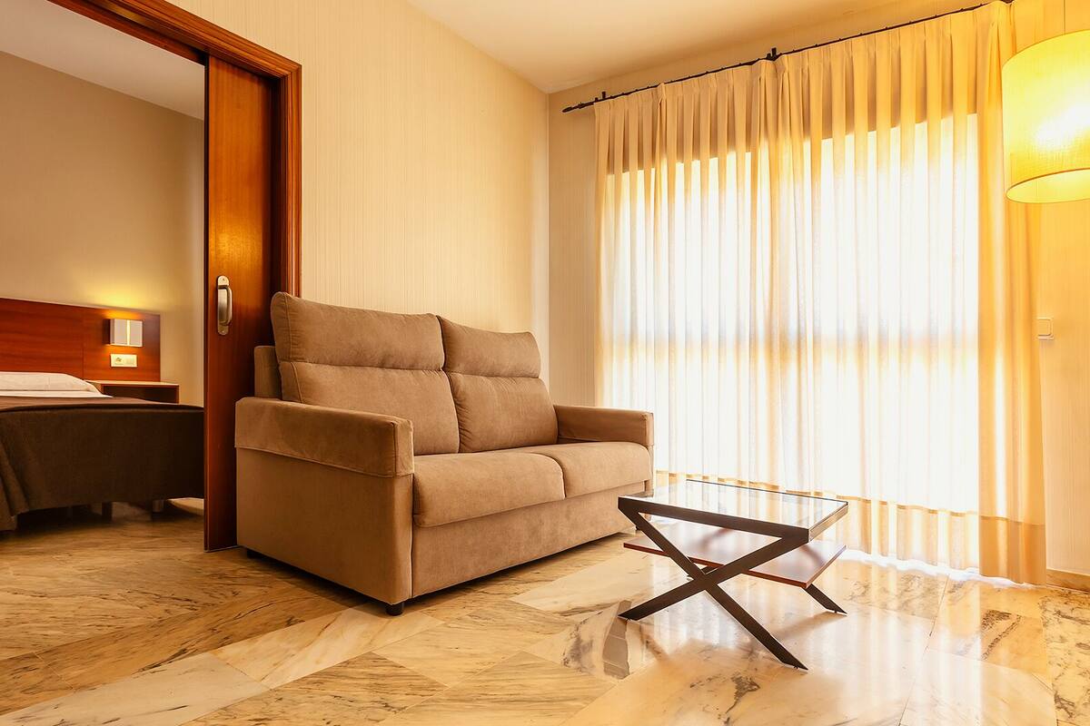 A hotel room with a sofa, a small table, a bed, and a window with curtains.