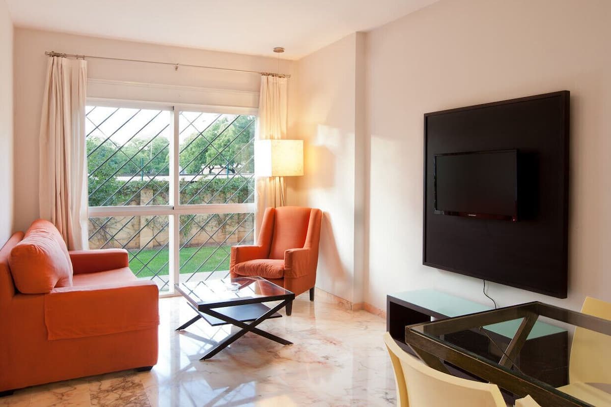 A modern living room with a sofa, armchair, and glass coffee table. A flat-screen TV is mounted on the wall, and a window with curtains provides a view of the outdoors.