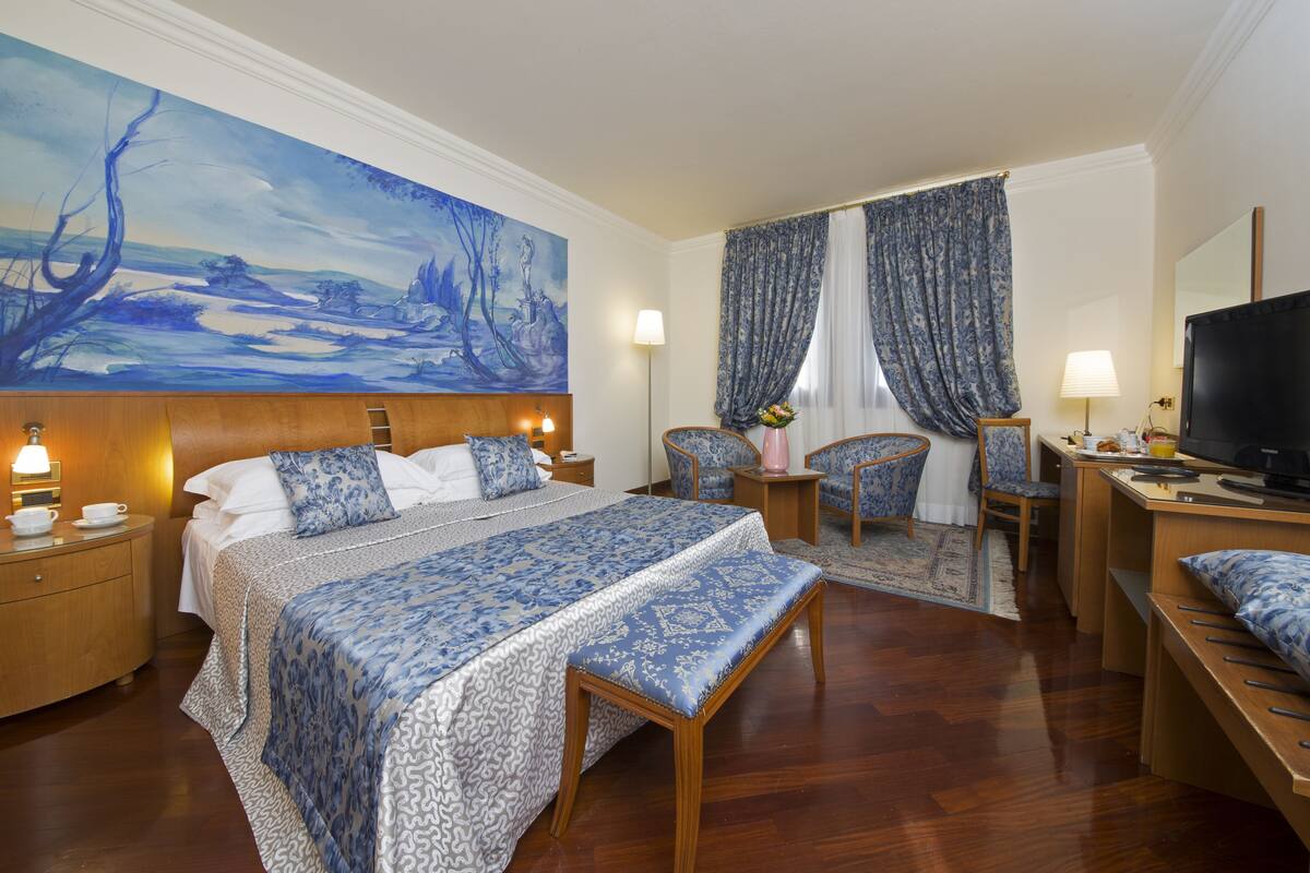 A hotel room with a large bed, a desk with a TV, a chair, a desk lamp, and a painting on the wall.
