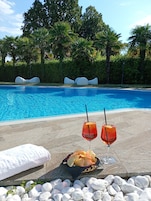 Zona de piscina con una piscina azul cristalina, dos bebidas con pajitas, una cesta de patatas fritas y una toalla blanca.