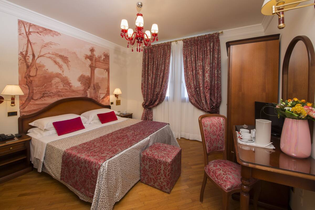 A hotel room with a large bed, a wooden nightstand, a desk with a chair, a mirror, and a chandelier.