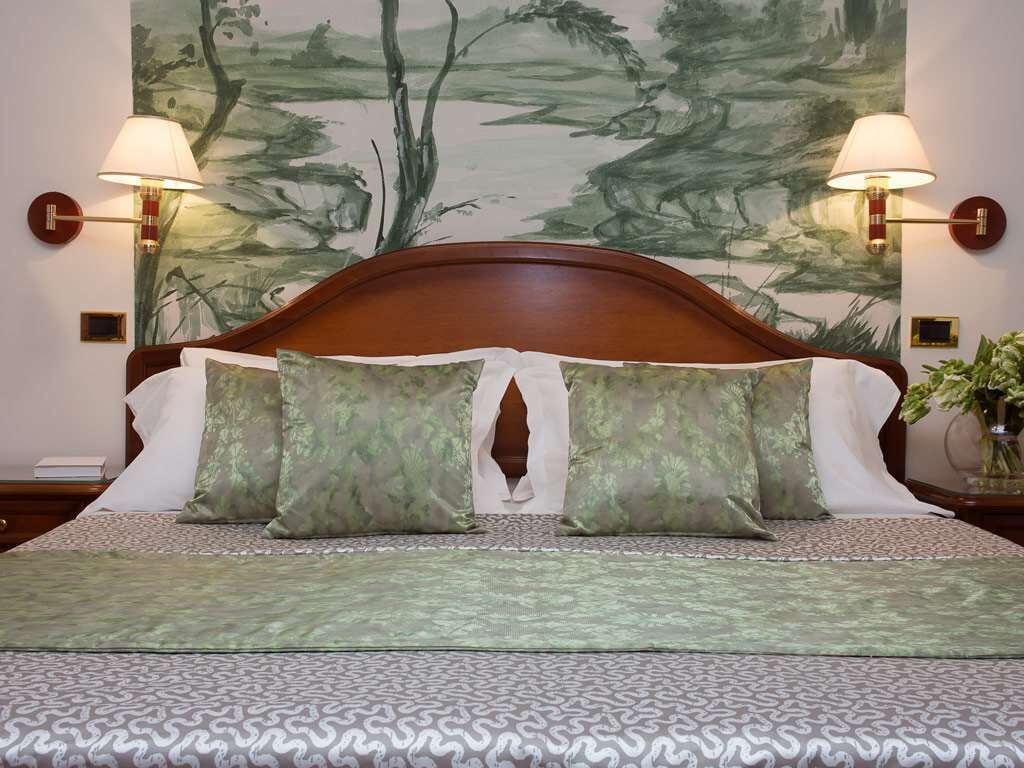 A neatly made bed with a patterned bedspread, two wall-mounted lamps, and a large abstract painting on the wall behind the bed.
