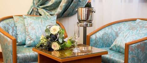 Una habitación con dos sillones tapizados, una mesita de madera con un arreglo floral y una botella de champán, y un objeto metálico sobre la mesa.