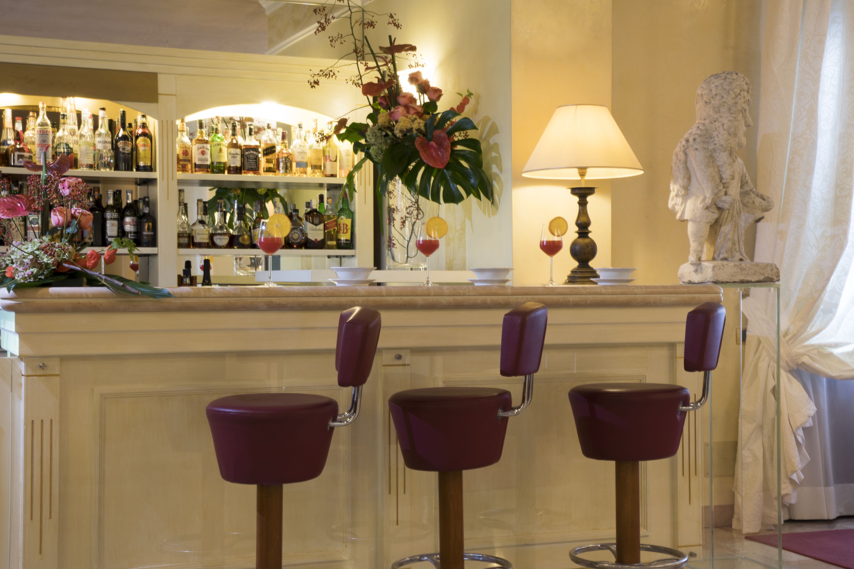 A bar area with three purple bar stools, a variety of liquor bottles on shelves, a decorative plant, and a statue.