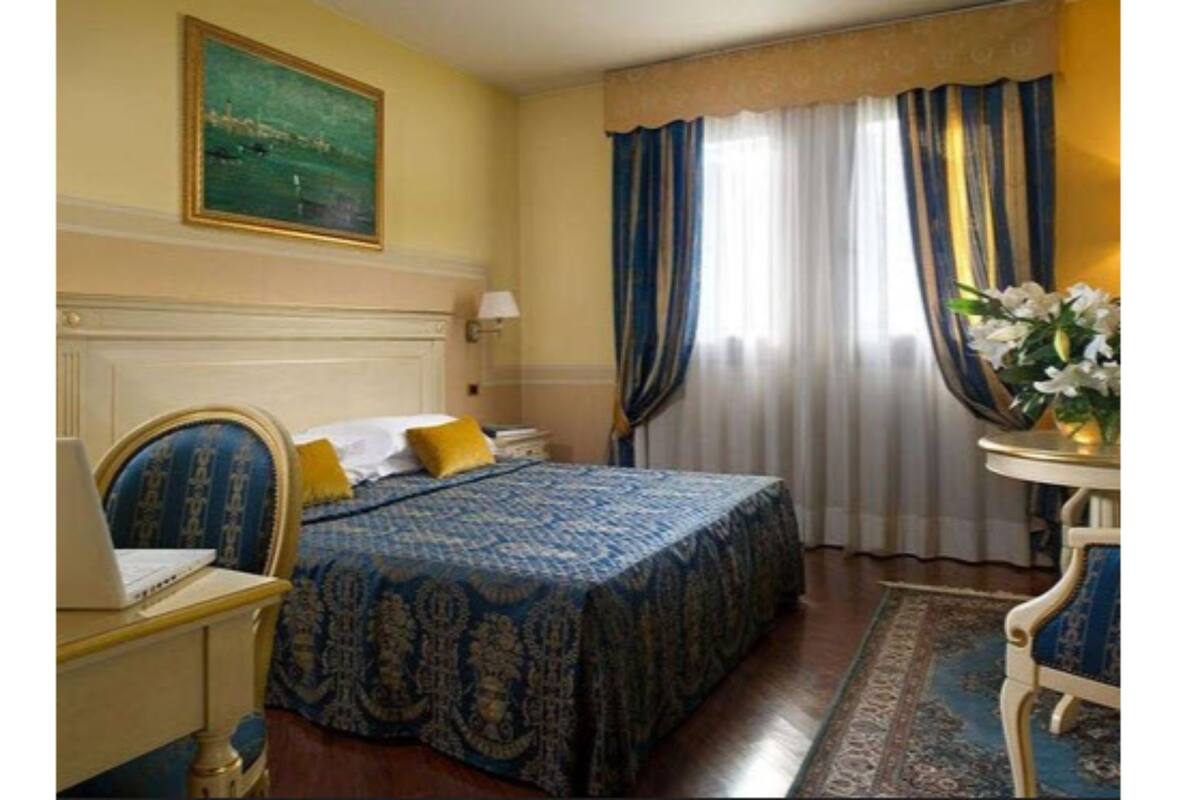 A hotel room with a bed, a desk with a laptop, a nightstand, a lamp, a window with curtains, and a painting on the wall.
