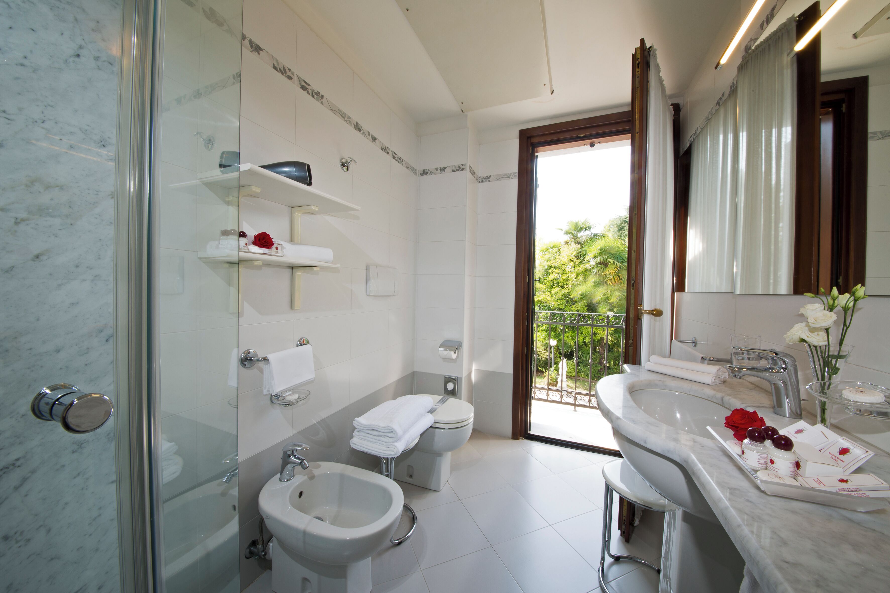 Un baño moderno con ducha envuelta en vidrio, un bidé, un lavamanos y un inodoro. Hay estantes blancos con artículos de tocador, un tocador con espejo y vistas a zonas verdes a través de la puerta.