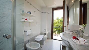 Un baño moderno con ducha envuelta en vidrio, un bidé, un lavamanos y un inodoro. Hay estantes blancos con artículos de tocador, un tocador con espejo y vistas a zonas verdes a través de la puerta.
