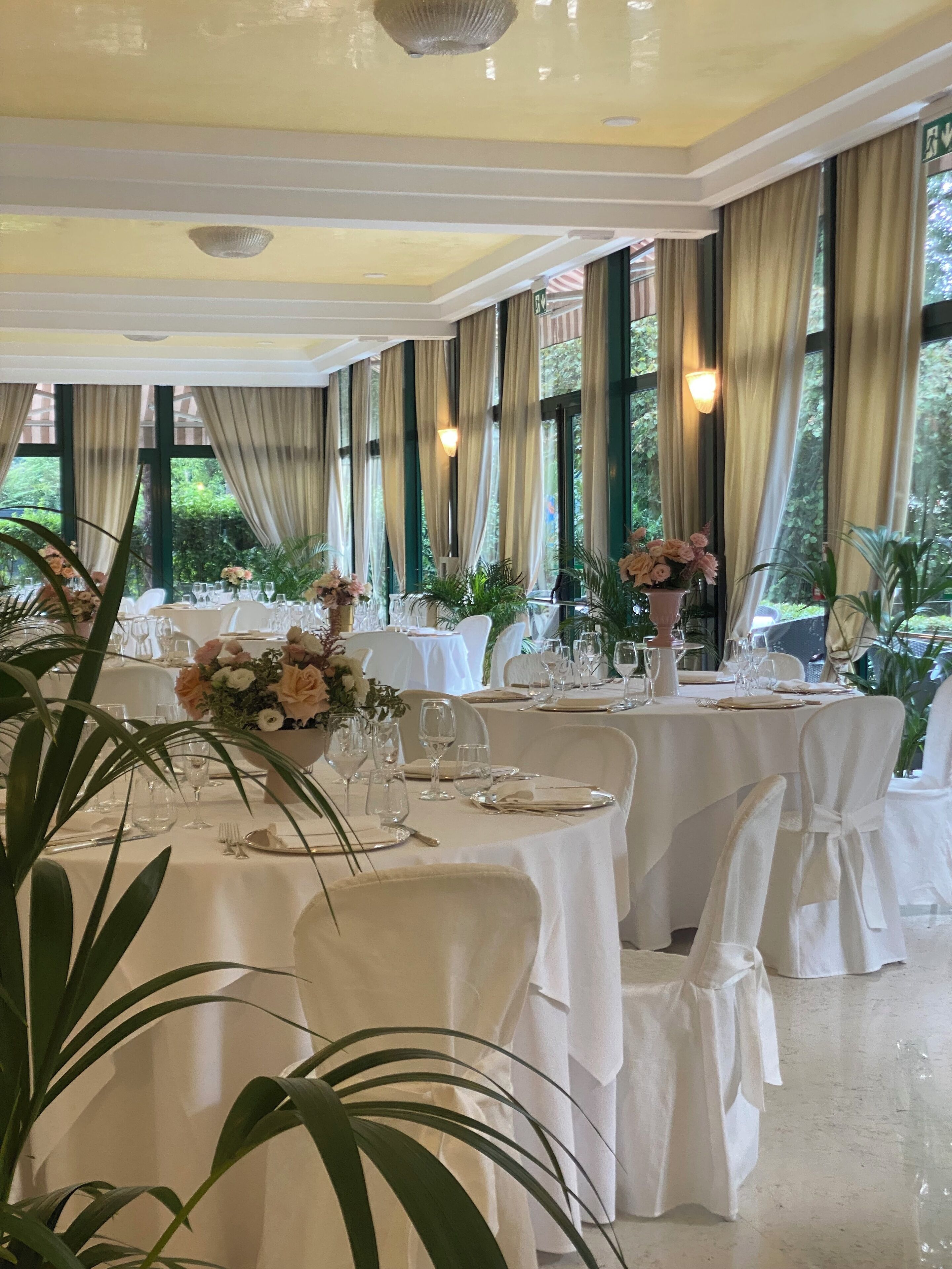 A banquet hall with round tables set for dining, large windows with curtains, and indoor plants.