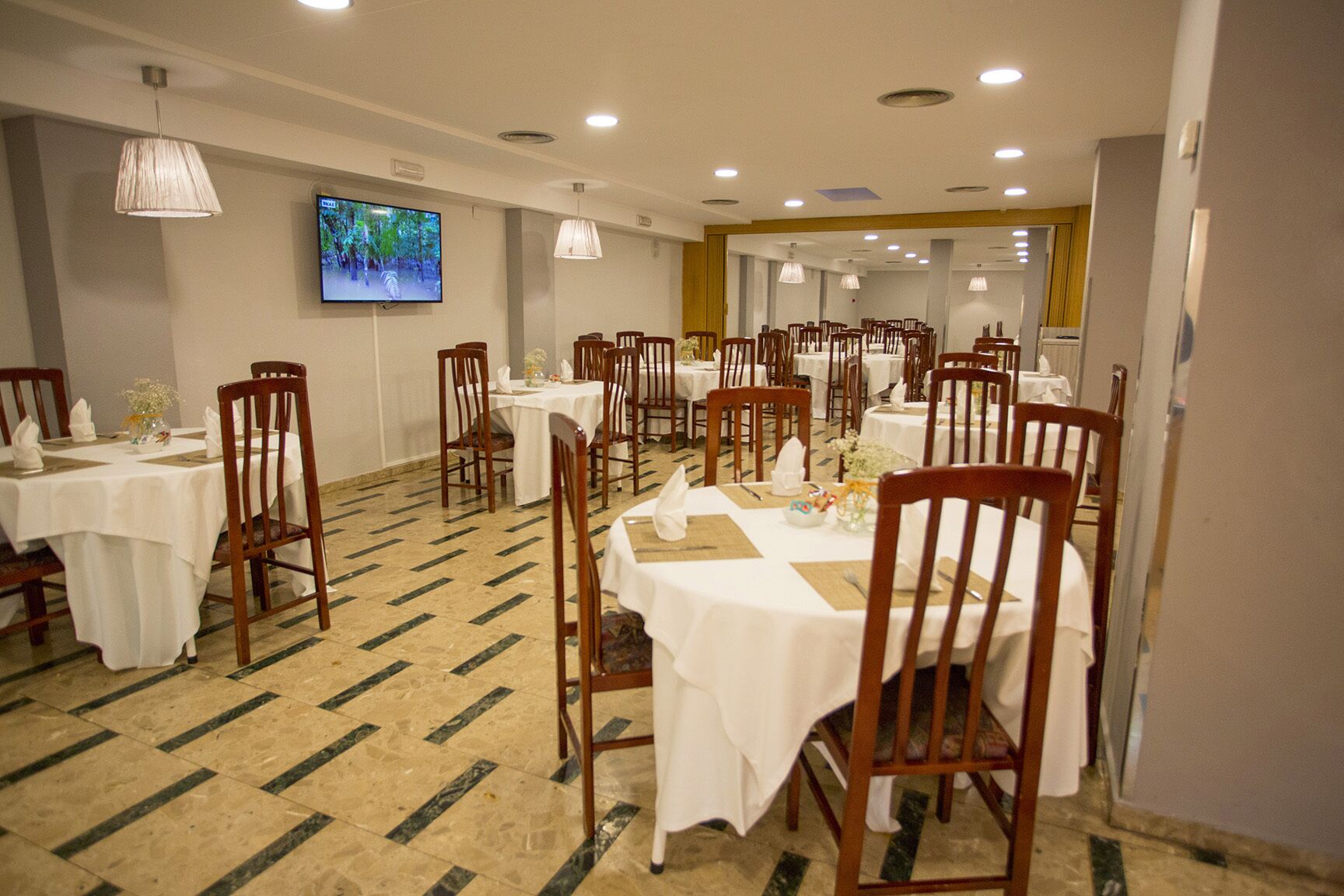 A dining area with multiple tables and chairs, a flat-screen TV on the wall, and patterned flooring.