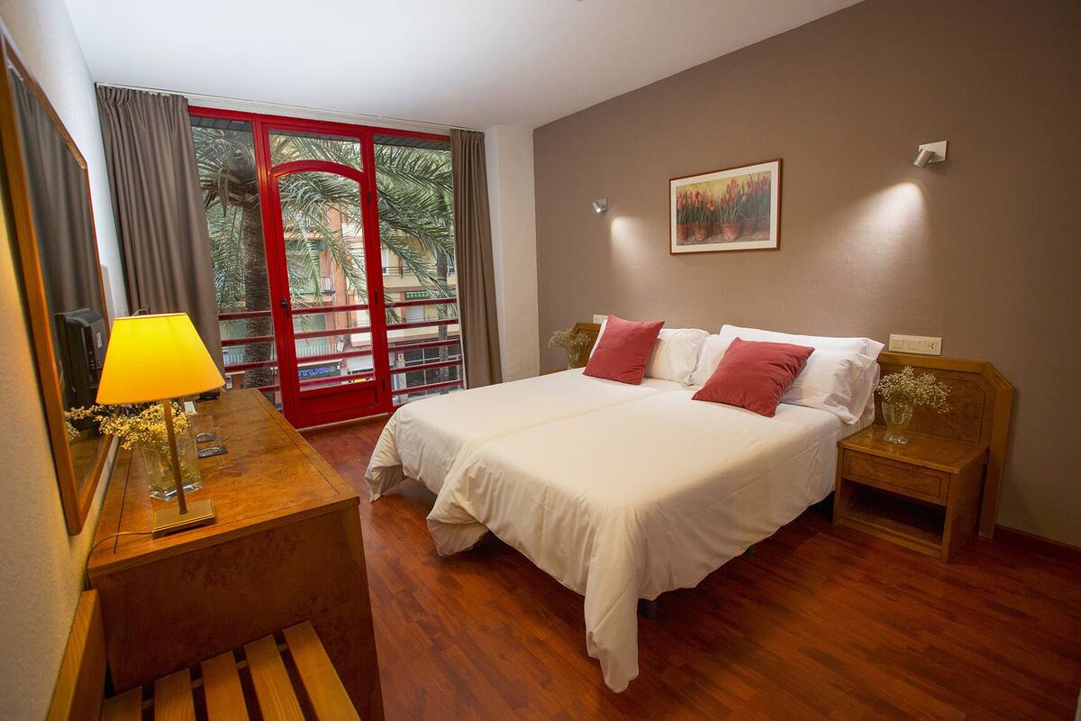 A hotel room with a bed, bedside tables, a lamp, a painting, and a view through a door.
