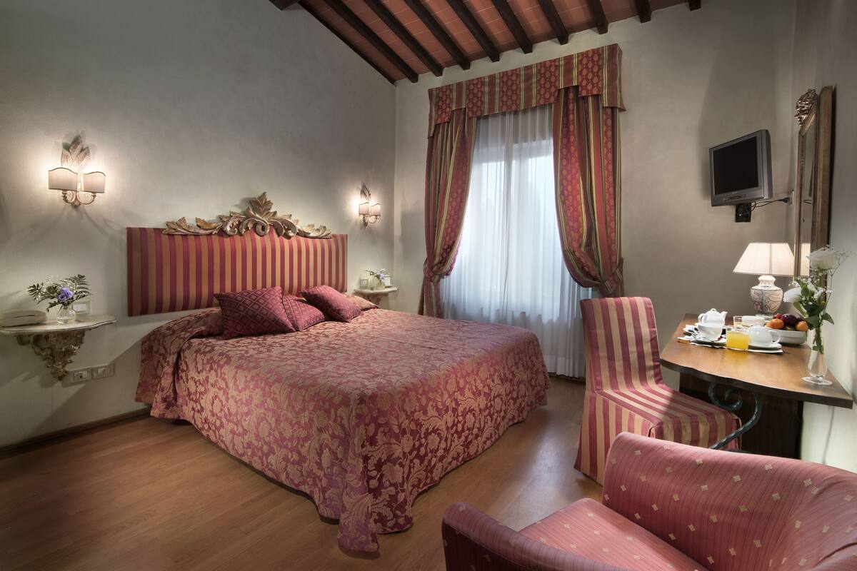 A hotel room with a large bed, a desk with a chair, a TV, and a bedside table with a lamp and flowers.