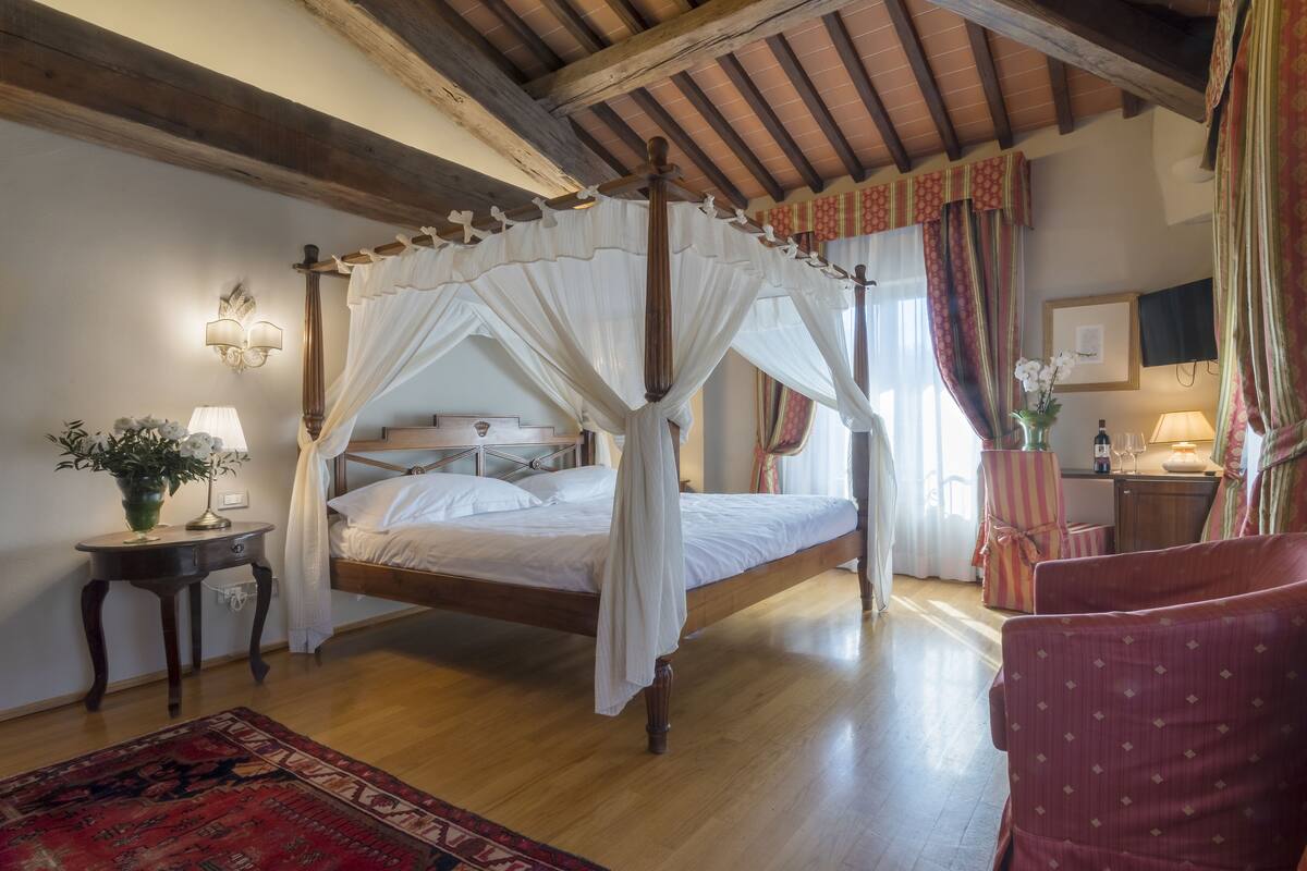 A four-poster canopy bed with a wooden frame, a bedside table with a lamp, and a red patterned rug.