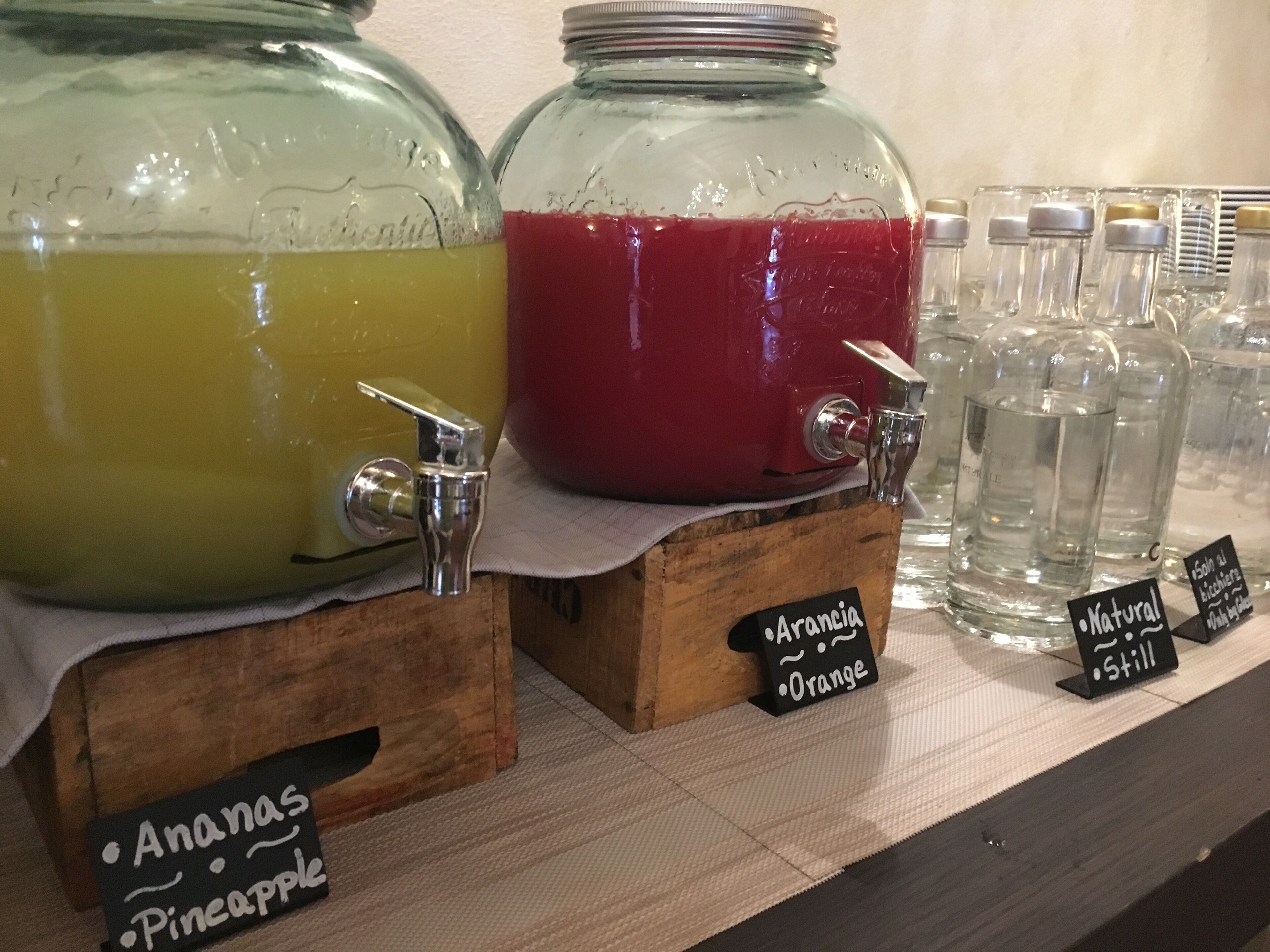 Deux grands bocaux en verre étiquetés avec différents noms de jus de fruits, un comptoir en bois et de plus petites bouteilles, également étiquetées.