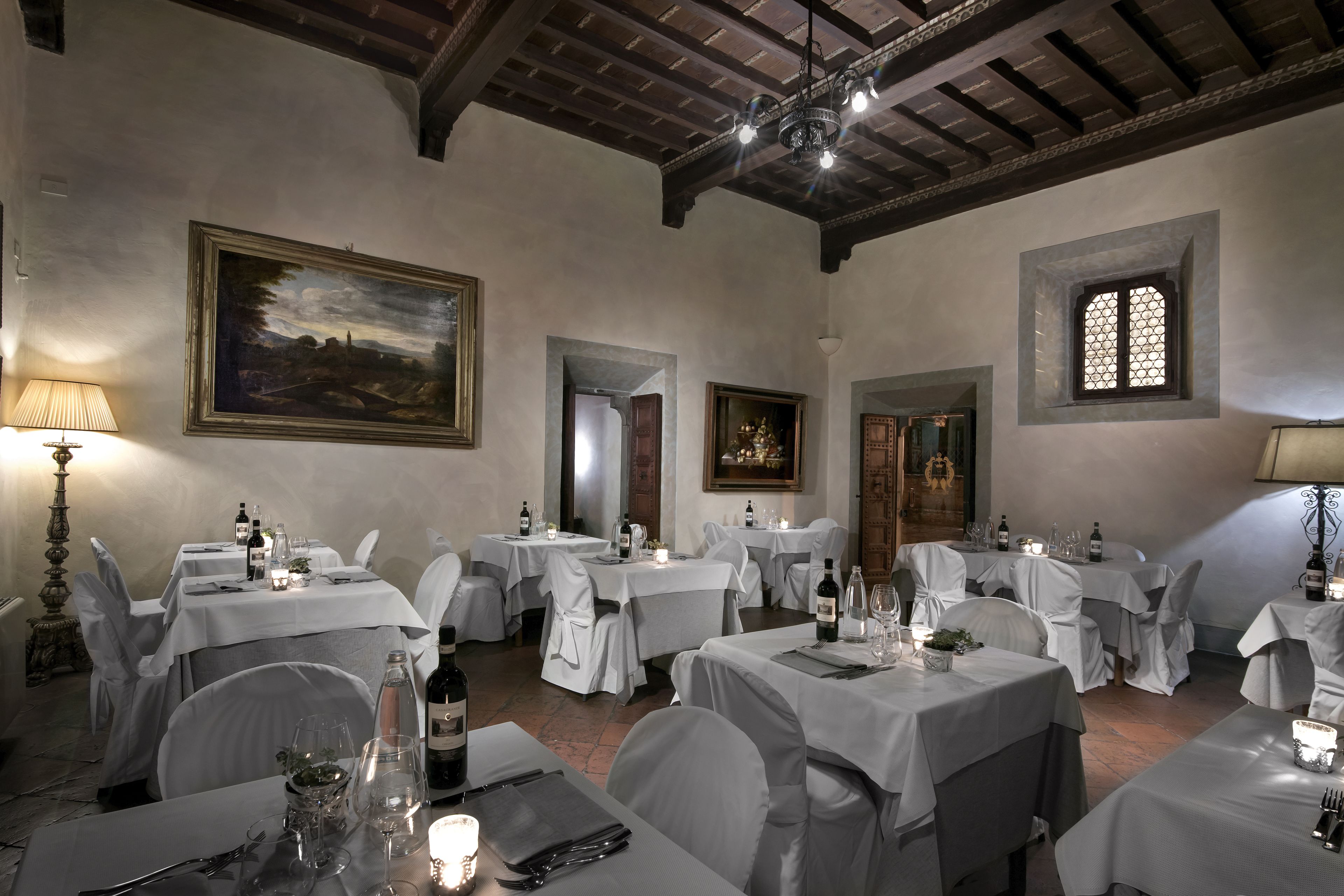 A dining area with multiple tables set for a meal, featuring white tablecloths and chairs, framed artwork on the walls, and a chandelier.