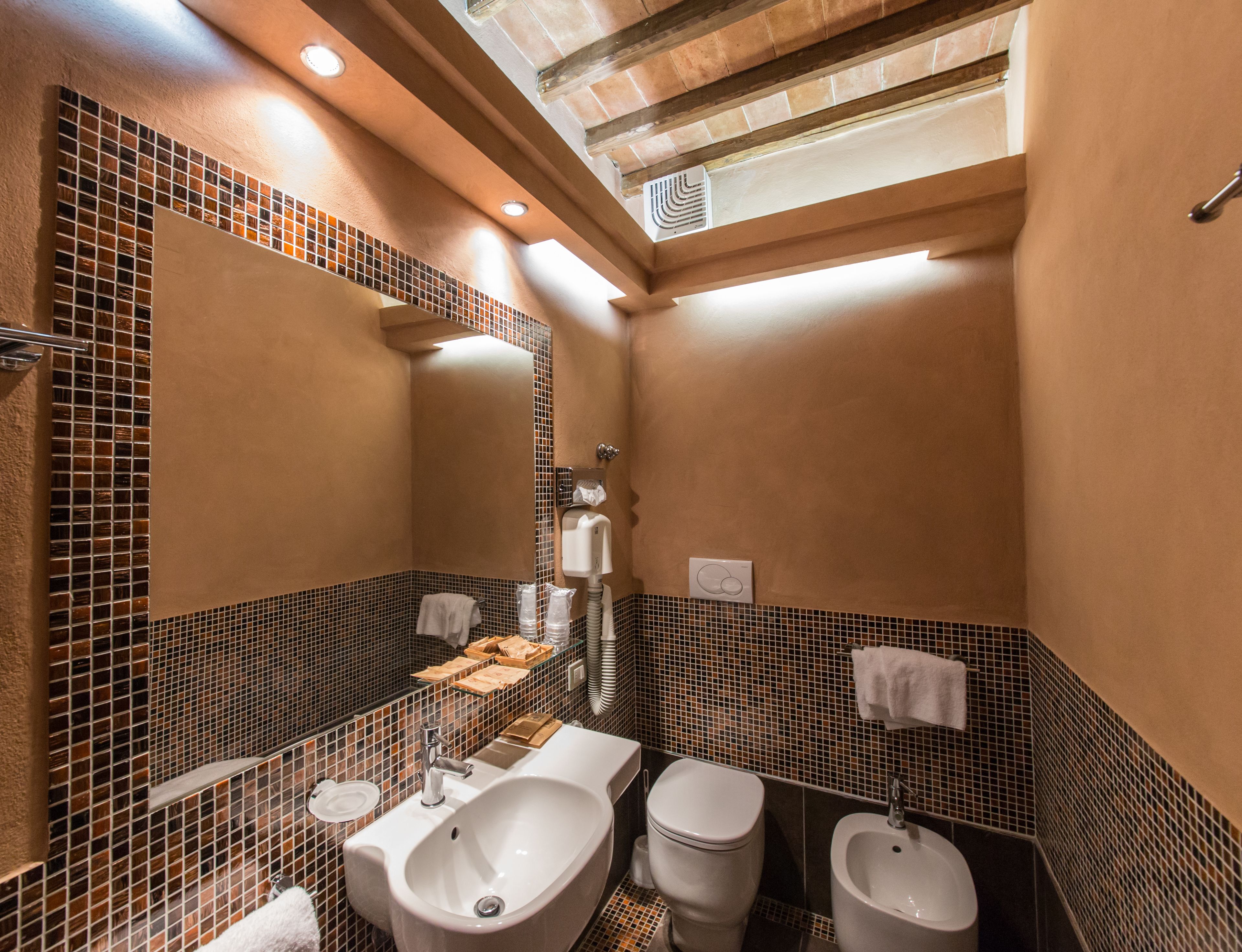 Ein Badezimmer mit einem großen Waschbecken, zwei Urinalen, einer Toilette und einem Bidet. Es gibt einen Spiegel, einen Handtuchhalter und ein Regal mit Pflegeprodukten.
