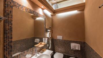 Un bagno con un lavandino grande, due orinatoi, un water e un bidet. Si nota uno specchio, un portasciugamani e uno scaffale con articoli da toilette.