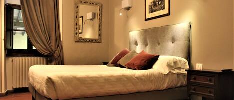A neatly made bed with a headboard, a bedside table, a nightstand, a framed picture on the wall, and a window with curtains.