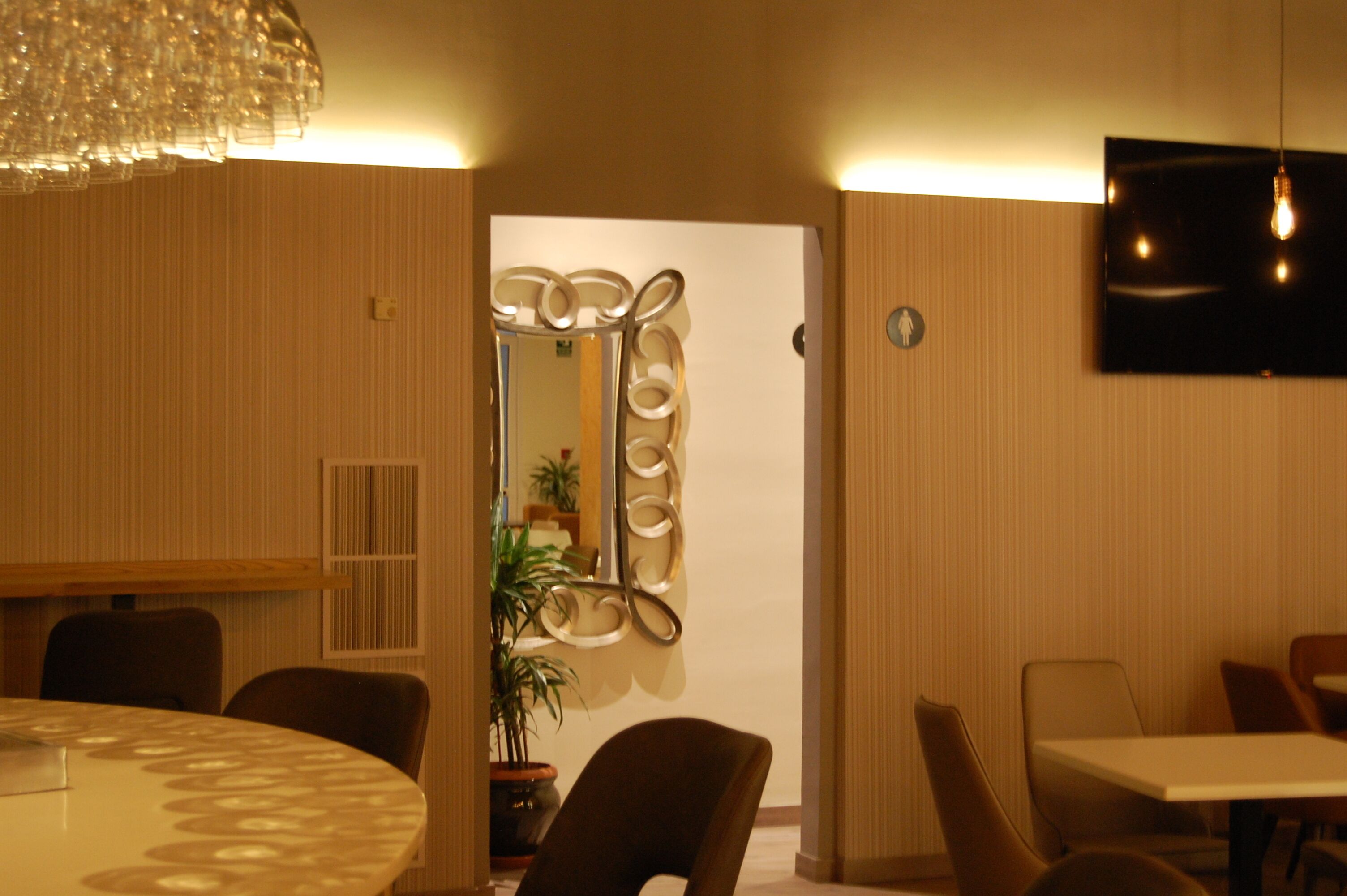 A hotel room with a round dining table, chairs, a potted plant, a decorative mirror, and a flat-screen TV mounted on the wall.