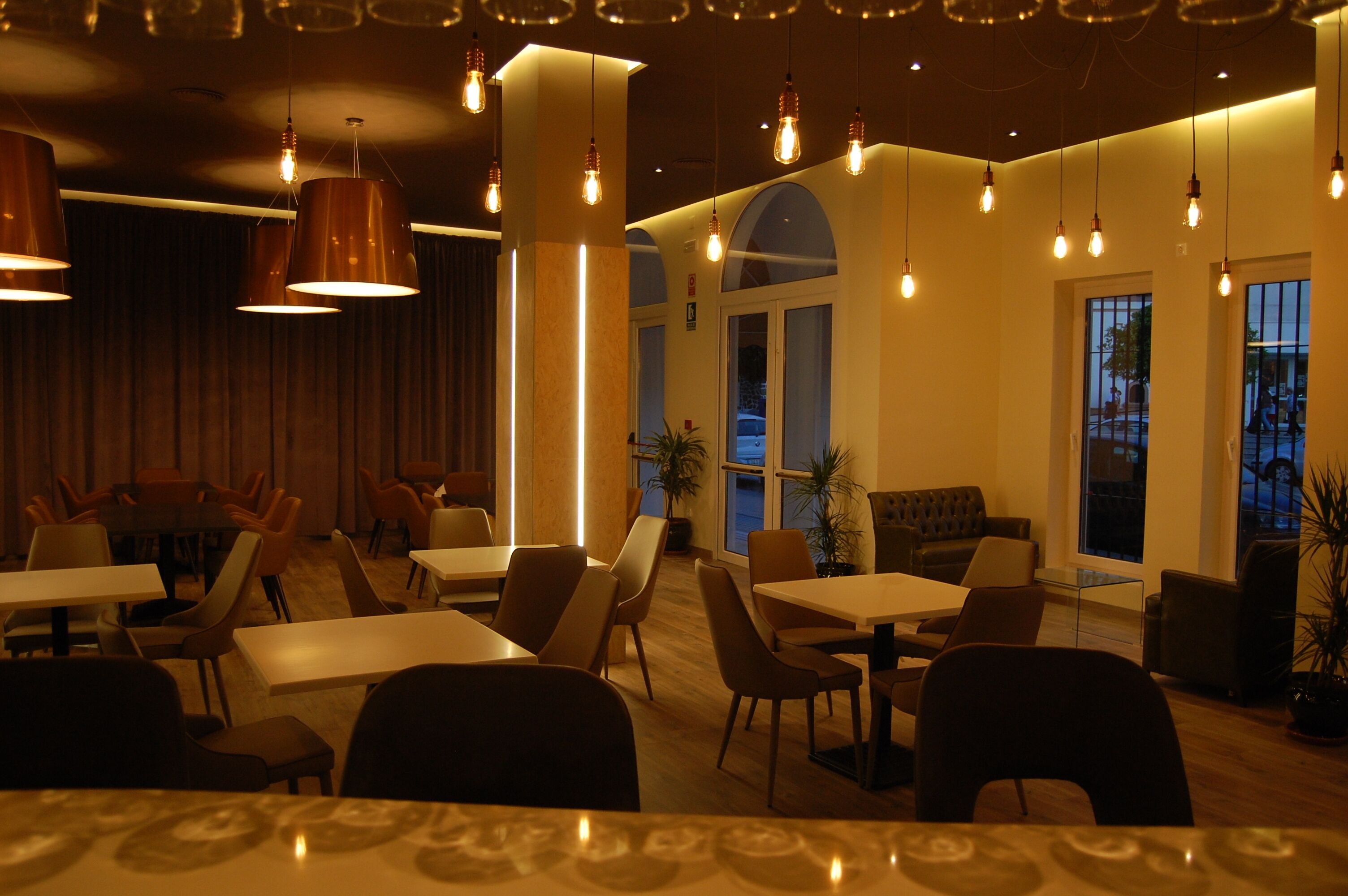 Een restaurant met tafels en stoelen, hangende lampen en grote ramen.