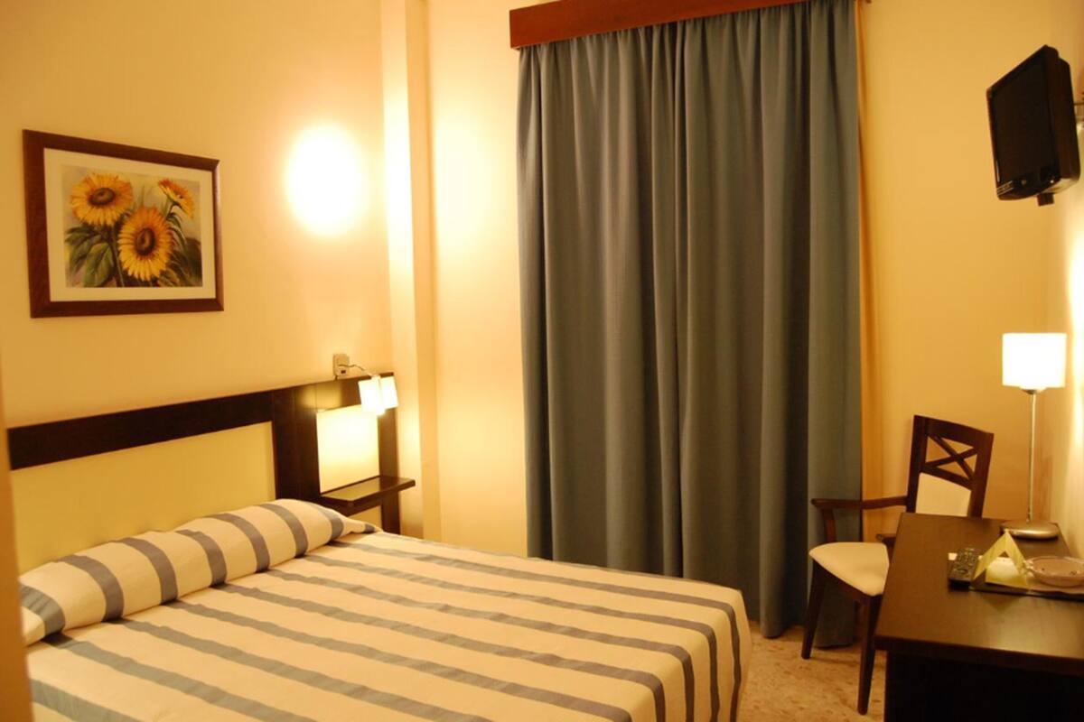A hotel room with a bed, a chair, a table, a lamp, a painting, and a curtain.