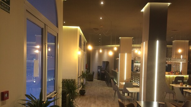 A modern restaurant with a long counter, potted plants, and a view of the city at night. - Hotel Castillo (Palma del Rio)